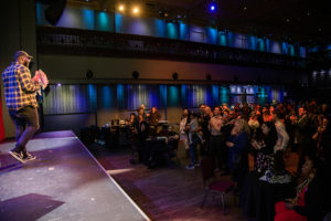 A Black man in a black and white flannel shirt, black jeans, black Converse sneakers and a black hat speaks from a raised platform to a crowd of individuals standing in a dimly lit room.