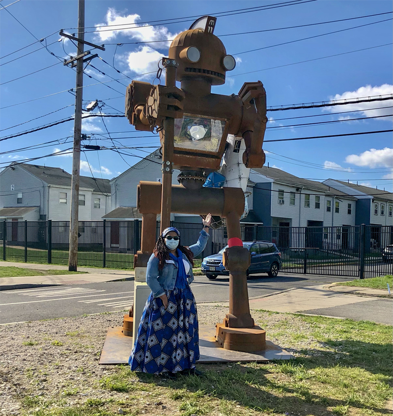 Photo of a Black woman standing in front of a large robot-esque-looking sculpture. The woman is wearing dark sunglasses, a blue medical mask, a blue head band over deadlocks, a denim jacket, and a blue skirt with white flowers. Behind the woman and sculpture are power lines, a blue SUV, a black fence enclosing a housing complex, and a blue sky with a few white clouds.
