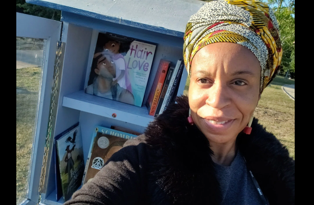 Photo of a Black woman with a yellow, white, black, and red head wrap standing in front of a blue hutch that has books inside. The front cover of the book "Hair Love," featuring a cartoon depction of a Black man and little girl looking at one another, is visible. 