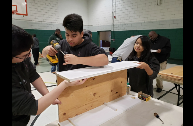 Photo of three teens putting together a wooden box that will be used for Little Free Library boxes. They are in what seems to be a gymnasium that is painted white on the top half and a dark green below. There is a white and red basketball net visible in the upper left hand portion of the photo. 