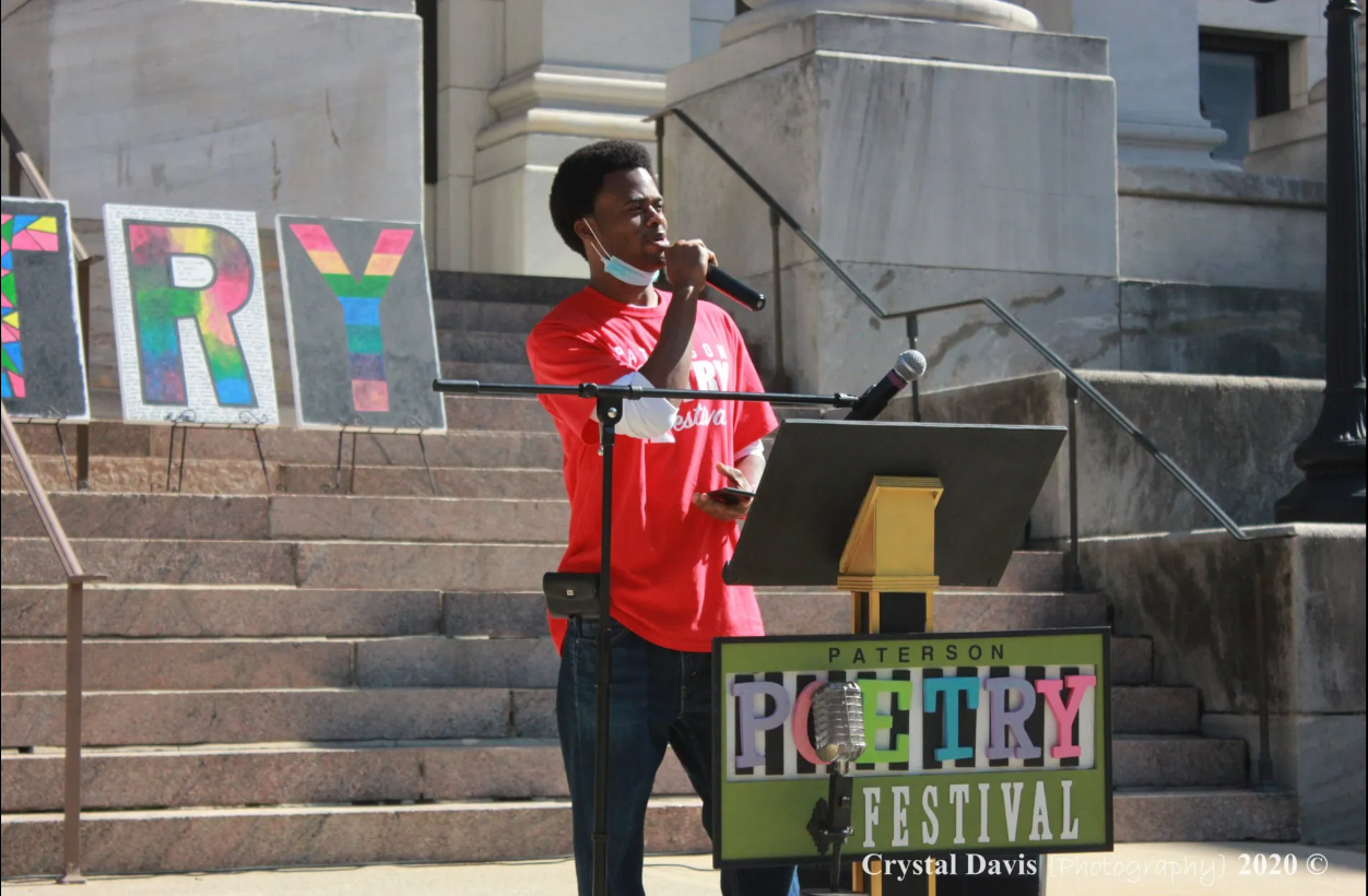 Photo of a young Black man in a salmon-colored tshirt is holding a microphone and speaking from behind a pedastol with a plackard that says Paterson Poetry Festival. The letters TRY are displayed behind him in rainbow colors on top of tan stone steps.