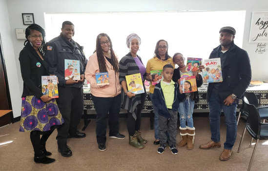 Photo of six Black adults and two Black children holding books and smiling for the camera in front of a table that has a black and white table cloth in front of a large window. The room they are in has white walls with a few black frames that seem to house word art on the walls. There are two black chairs to the right of the image, next to one of the adults. 